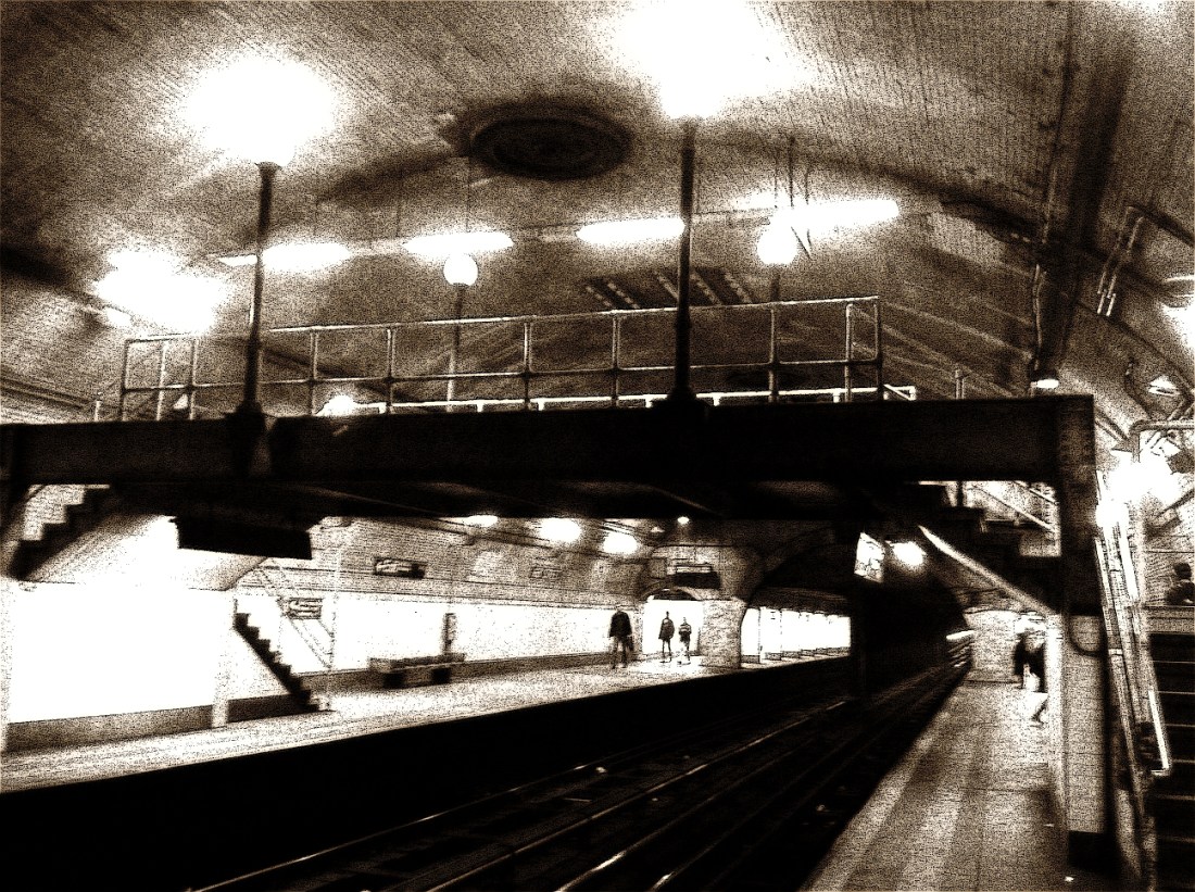 Black-and-white, sepia-toned photo of an old vaulted subway station with a steel footbridge over the tracks and a few figures on the platform.