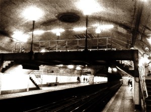 Black-and-white, sepia-toned photo of an old vaulted subway station with a steel footbridge over the tracks and a few figures on the platform.