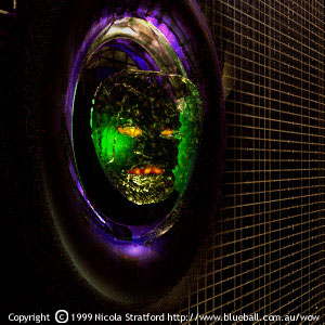 Dark digital image of a glowing green, mask-like alien face inside a purple-rimmed oval porthole set in a wall of fine gold grid lines