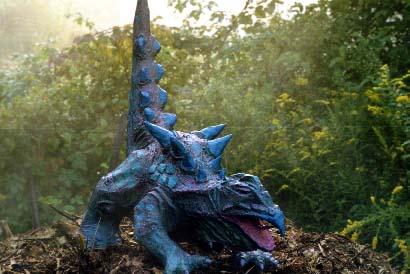 Photograph of a blue papier-mâché dragon-like creature with spines down its back and a pink tongue, sitting outdoors among green plants